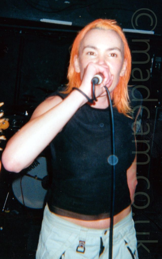 A woman with shoulder length orange hair, wearing a black t-shirt and grey trousers, holding a microphone to her mouth, with the cord for the mic wrapped around her right wrist. She is staring past the camera. A drum kit is visible in the background, with black walls and ceiling filling the rest of the frame.