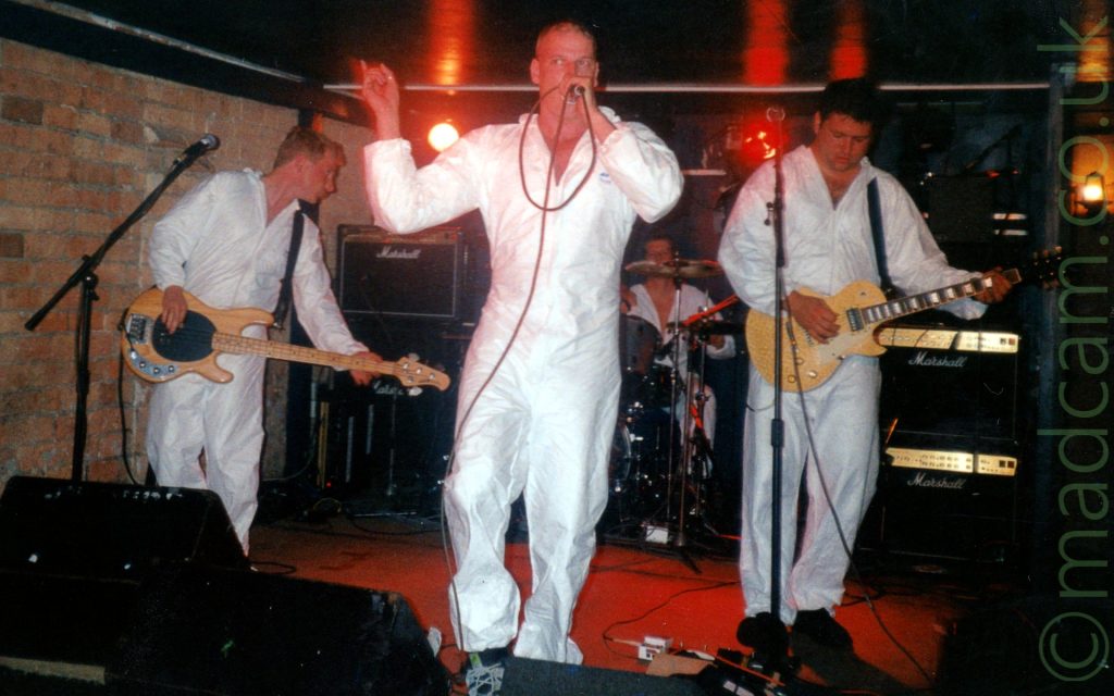 4 members of a band performing on stage, all wearing white coveralls. On the left is a man with short dark hair, playing a brwon bass guitar, and looking towards the right of the frame. In the middle is a man with a shaven head, holding a microphone to his mouth with his left hand, his right hand raised into the air. At the back is a man sitting behind a black drum kit. On the right is a man with short dark hair, playing a brown electric guitar, and staring at the red stage floor. A brown brick wall is on the left of the background, the rest of the walls and ceiling being black. Red lights on a rack at the rear of the stage add some dramatic lighting.