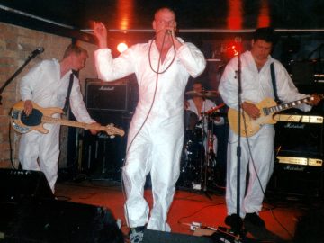 4 members of a band performing on stage, all wearing white coveralls. On the left is a man with short dark hair, playing a brwon bass guitar, and looking towards the right of the frame. In the middle is a man with a shaven head, holding a microphone to his mouth with his left hand, his right hand raised into the air. At the back is a man sitting behind a black drum kit. On the right is a man with short dark hair, playing a brown electric guitar, and staring at the red stage floor. A brown brick wall is on the left of the background, the rest of the walls and ceiling being black. Red lights on a rack at the rear of the stage add some dramatic lighting.