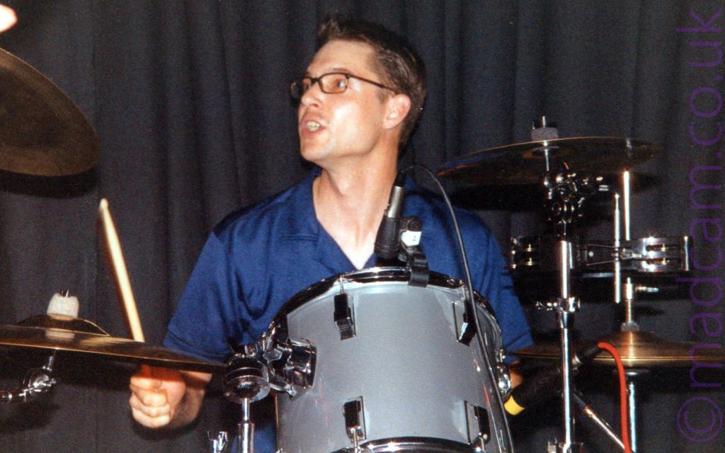 A man with short dark hair, wearing a pair of dark-rimmed glasses and a shiny blue shirt, sitting behind a grey drumkit, with a drumstick raised in his right hand, as looks off the left of the frame. A black curtain fills the background behind him.