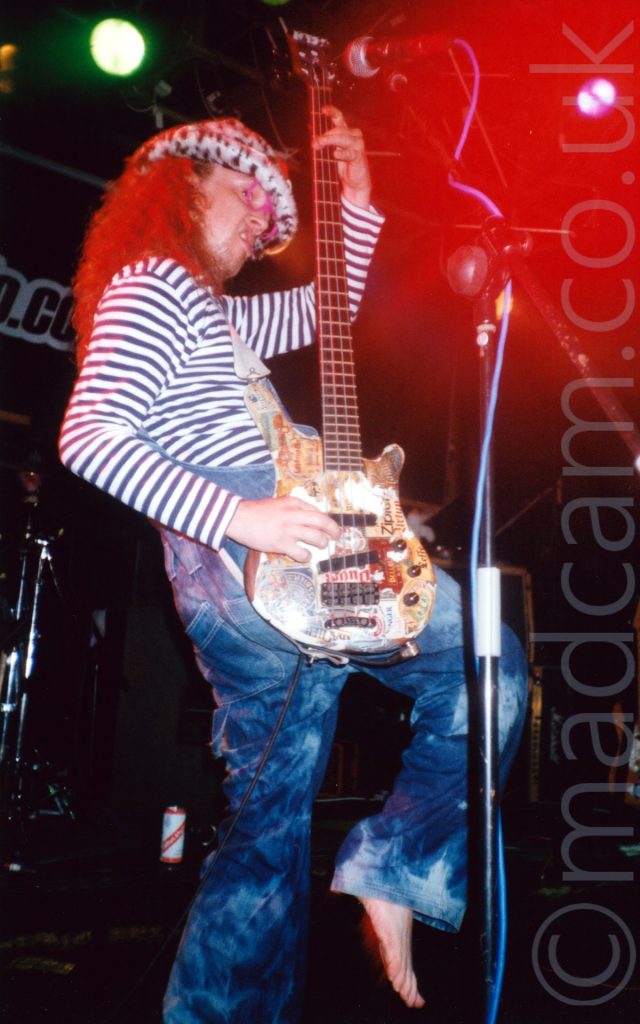 A man with long brown hair cascading over his shoulders facing to the right, wearing a furry, black and white animal-print hat, black and white striped long-sleeved t-shirt and blue denim dungarees, playing a bass guitar that he is holding vertically, while snarling and raising his right foot off the ground, his knee raised high in front of him. A red light is shining on his hair, with another red light in the top right corner creating a bloom of light around his microphone. Black walls fill the rest of the background.