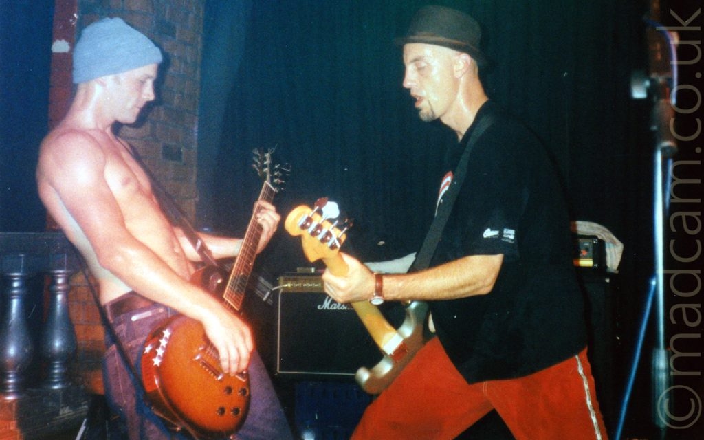 2 men performing on a stage as part of a band. On the left is a rather sweaty man, inexplicably wearing a grey wollen hat and no top, with a blue pair of jeans, and playing a brown electric guitar. On the right is a tall man wearing a black bowler hat, a black t-shirt, and a red pair of trousers with a silver stripe running down the side of the leg, playing a silvery-grey bass guitar. A grey balustrade can be seen off to the left of the frame, next to a brown brick wall, with black walls filling the rest of the background.