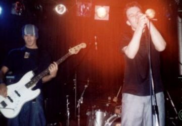 2 men on a stage, performing as part of a band. On the let is a tall man wearing a blue, grey and white woolly hat, dark t-shirt, and blue jeans, and playing a white bass guitar. On the right is a man with short dark hair, wearing a dark shirt and grey trousers, holding a microphone in both hands, even though it's on a stand, while he sings in to it. In the background, a man can almost be seen sitting behind a silver drum kit, only his right hand visible clutching a drum stick. Black walls and bright stage lights fill the rest of the background, with a bloom of red light above the singers head.