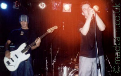 2 men on a stage, performing as part of a band. On the let is a tall man wearing a blue, grey and white woolly hat, dark t-shirt, and blue jeans, and playing a white bass guitar. On the right is a man with short dark hair, wearing a dark shirt and grey trousers, holding a microphone in both hands, even though it's on a stand, while he sings in to it. In the background, a man can almost be seen sitting behind a silver drum kit, only his right hand visible clutching a drum stick. Black walls and bright stage lights fill the rest of the background, with a bloom of red light above the singers head.