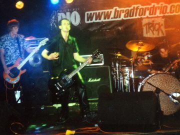 3 people on stage, performing as part of a rock band. On the left is a man with shaggy, ear-length brown hair, wearing a blue and grey shirt, and playing an orange bass guitar. In the middle is a man with short dark hair, wearing a black shirt and dark trousers, playing a black electric guitar. On the right is a man with short dark hair, sitting behind (and playing) a black drum kit at the back of the stage. The wall at the back is black, with large black text "www,bradfordrio.com" outlined in white, with a smaller, white sign below with the black word "Trick". There are different coloured lights aimed at each person on stage - the bassist is highlighted in blue, an acid green glow surrounds the guitarist while a bloom of orange light surrounds the drummer.
