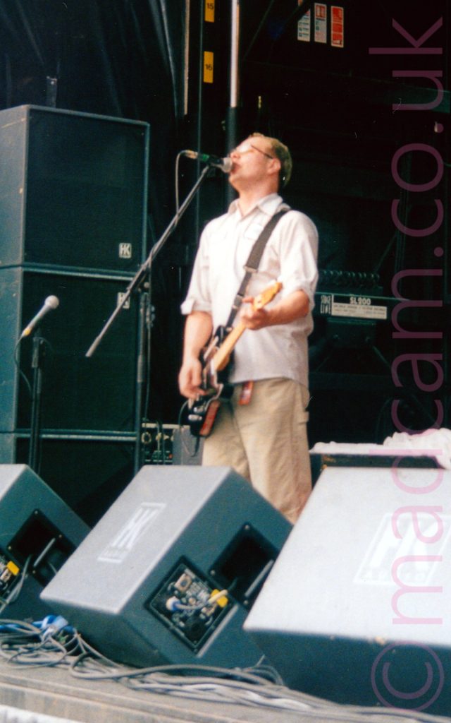 A man with short brown hair, wearing a white shirt and khaki pants, playing a black electric guitar and singing into a microphone on a stand in front of him on an open air stage. The mic stand is obviously set up for someone a little bit taller than him! Monitor speakers litter the front of the stage, with vertical stacks of speakers and amps in the background.