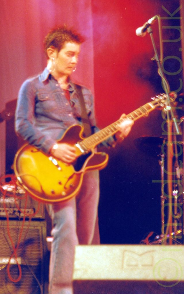 A woman with short, spiky brown hair, wearing a blue denim shirt over a red t-shirt and a grey pair of trousers, playing a yellow and brown electric guitar, and looking down at her left hand. A microphone on a stand looms over the right of the frame, while stacks of amps are on the right in the background. A white curtain, coloured red by the stage lights, fills the rest of the frame.