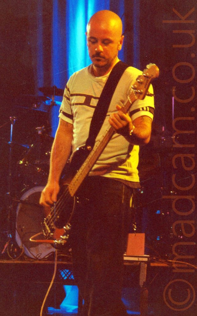 A man with a shaven head, wearing a white t-shirt with a black bar across the chest and dark trousers, playing a bass guitar, and looking down rather pensively, performing on a stage. A drumkit on a riser is in the background behind him, with a curtain as a backdrop, the centre section illuminated blue by the stage lights.