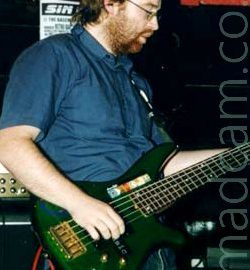 A man with ear-length brown hair and a short brown beard, wearing a blue shirt and a dirty pair of blue jeans, playing a green 5-string bass guitar, facing to the right. Several amplifiers can be seen in the background in front of red and black walls.