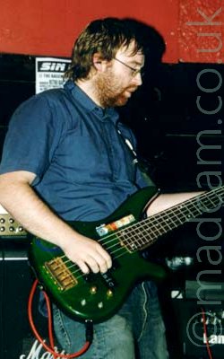 A man with ear-length brown hair and a short brown beard, wearing a blue shirt and a dirty pair of blue jeans, playing a green 5-string bass guitar, facing to the right. Several amplifiers can be seen in the background in front of red and black walls.