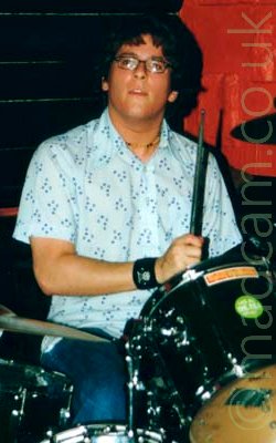 A man with fairly short dark hair, wearing a pair of eye-glasses, a white shirt with blue dots, and a pair of blue jeans, sitting behind a black drum kit, a stick raised in his right hand, as he stares off into the distance past the cameras left. red and black walls fill the background.