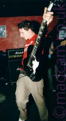 A man with short dark hair, wearing a red t-shirt and beige trousers, holding a black electric guitar aloft by the neck in his left hand, his right hand nowhere to be seen standing almost side-on to the camera, facing to the left of the frame. Amplifiers and speakers line the red and black walls in the background.