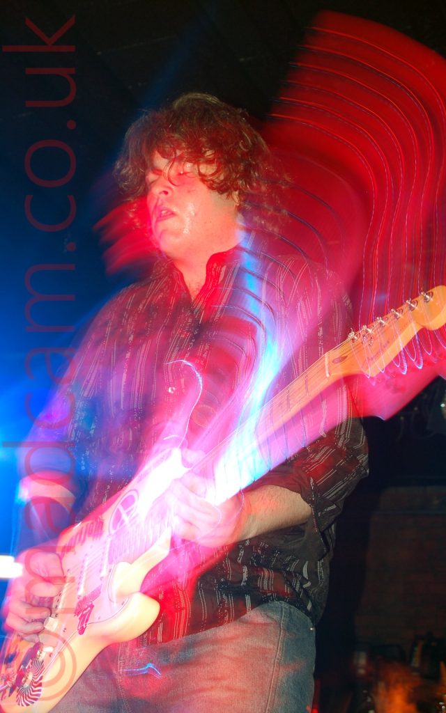A man, with mid-length shaggy hair, wearing a stripy brown shirt and a pair of jeans, playing a yellow electric guitar with his eyes shut. A long exposure and his movement has left red trails of light with the occasional blue streak as the neck of his guitar moves through an arc, another red streak by his head shows where that has moved. Black walls in the background fill the rest of the frame, apart from by his right elbows, where an explosion of blue light illuminates that part of the frame.