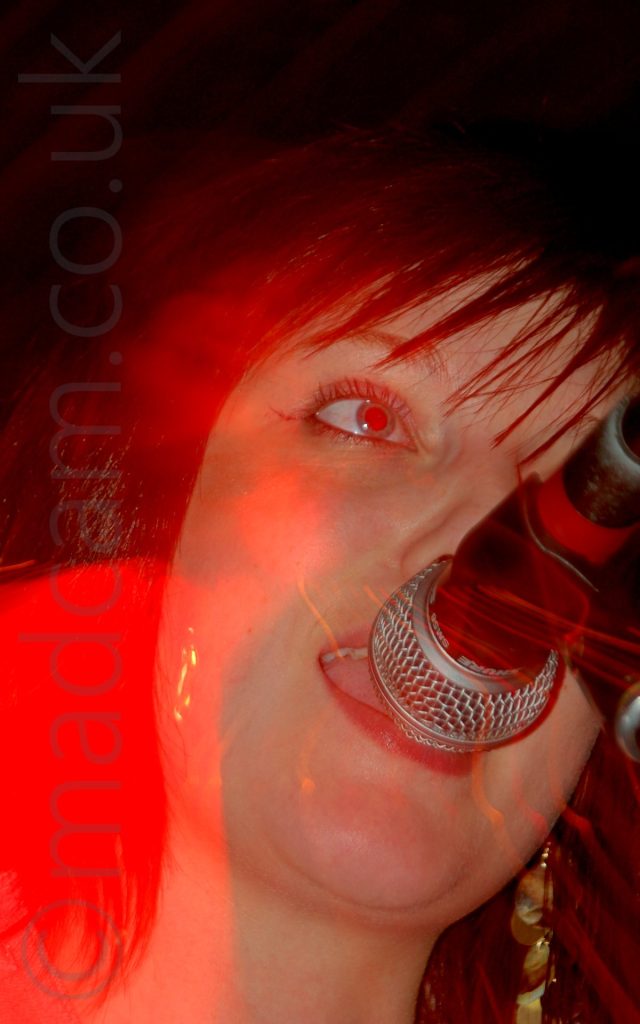 Close-up of the face of a woman as sings into a microphone in front of her mouth, staring off into the distance. A bloom of red light illuminates the right side of her face, with black walls and ceiling filling the rest of the frame.