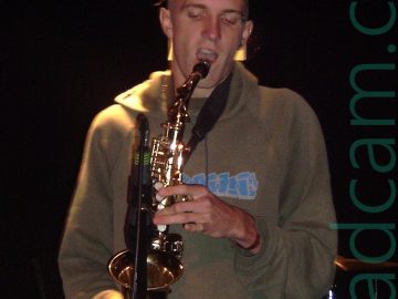 A man with a long, spiky but slightly wilting brown mohawk hairstyle, wearing a grey hoodie with some indecipherable blue text on the front, and a black pair of trousers. He is holding a saxophone up to an inverted microphone on a stand in front of him, and is opening his mouth to take a breath before playing it. A bright light is shining down on his right hand and shoulder. Black walls and ceiling fill the background, with a slightly illuminated heating vent right behind his head.
