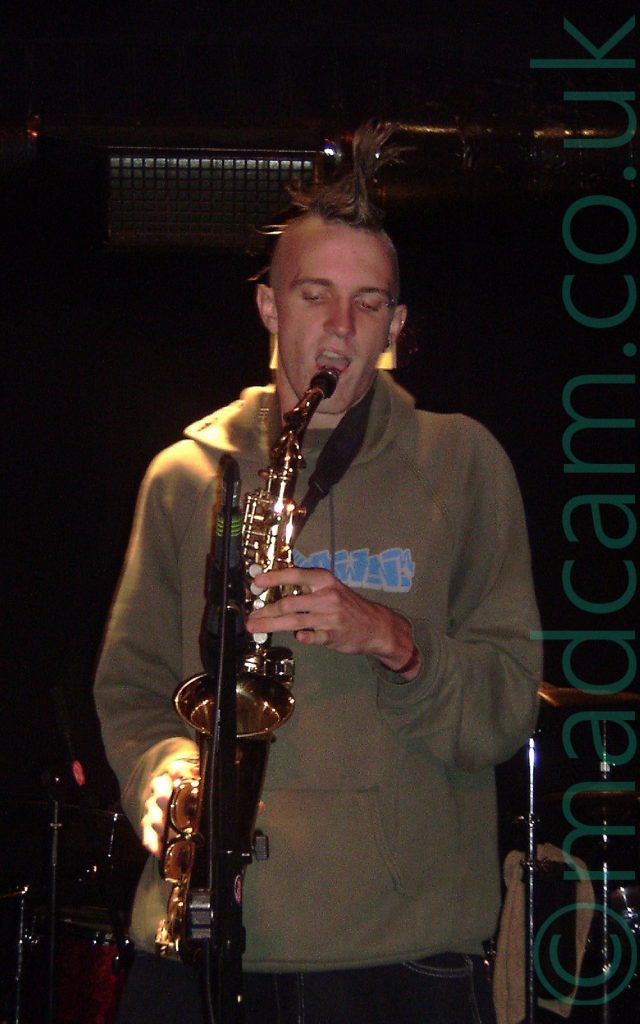 A man with a long, spiky but slightly wilting brown mohawk hairstyle, wearing a grey hoodie with some indecipherable blue text on the front, and a black pair of trousers. He is holding a saxophone up to an inverted microphone on a stand in front of him, and is opening his mouth to take a breath before playing it. A bright light is shining down on his right hand and shoulder. Black walls and ceiling fill the background, with a slightly illuminated heating vent right behind his head.