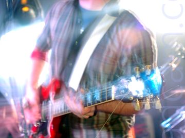 A man with short dark hair and beard, wearing a white shirt with broad red and blue vertical stripes over a grey t-shirt, and a blue pair of jeans, playing a red electric guitar, and screaming into a microphone on a stand in front of him. A long exposure, combined with his movement and the stage lights has resulted in some blurring of the image, with explosions of white light around him, reflecting off his guitar and face.