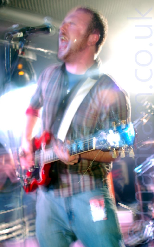 A man with short dark hair and beard, wearing a white shirt with broad red and blue vertical stripes over a grey t-shirt, and a blue pair of jeans, playing a red electric guitar, and screaming into a microphone on a stand in front of him. A long exposure, combined with his movement and the stage lights has resulted in some blurring of the image, with explosions of white light around him, reflecting off his guitar and face.