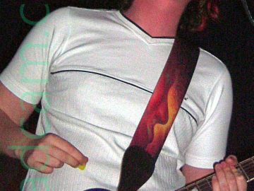 A man performing on a stage as part of a rock band. He has floppy long hair, and is wearing a white t-shirt over blue jeans, and has a blue electric guitar slung around his neck on a long black strap. He is singing in to a microphone on a stand in front of him as he plays his guitar. Black walls and ceiling fill the rest of the frame.