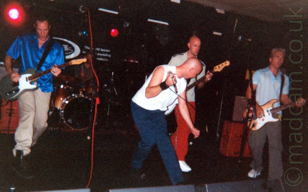 5 members of a band performing on a stage. On the left is a man with short, dark hair, wearing a shiny blue shirt and beige trousers, and playing a grey and white electric guitar. At the back is a man with short dark hair, wearing a grey shirt, sitting behind a reddish-gold drumkit. In the middle of the frame is a man with a shaved head, wearing a white sleeveless t-shirt and blue trousers, with a leather wristband around his right wrist, and holding a microphone in his right hand. He is slightly crouching over, and looking over to his left. Next is a man with a shaved head, wearing a lime green t-shirt and red trousers, and playing a white bass guitar. On the right is a man with short, light-coloured hair, wearing a blue and white striped t-shirt and grey trousers, playing a brown and white electric guitar. Black walls and white ceiling fill the rest of the frame, with a pair of red stage lights on the upper left of the frame.