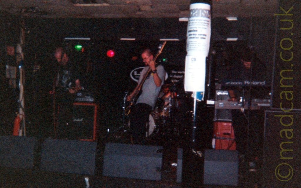 A rather dark and gloomy (and probably over-processed) photo of the 4 members of a band performing on stage. On the left is a man with a shaven head, a black t-shirt with a white logo, and dark trousers, playing an electric guitar. In the middle is a man with short dark hair, wearing a grey t-shirt and black trousers, and playing a bass guitar. Behind him is a man with short dark hair, wearing a white t-shirt, and sitting behind a drum kit. On the right is a man with short dark hair, wearing glasses, and a dark t-shirt and trousers, looking down at the bank of keyboards he is playing. A thick black pole dominates the foreground, slightly off to the right of centre, with some sheets of white paper taped to it, with only the word "Oceansize" discernible at the top. A row of grey monitor speakers line the front of the stage. Black walls and a grey ceiling fill the rest of the frame.