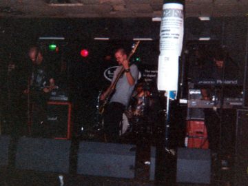 A rather dark and gloomy (and probably over-processed) photo of the 4 members of a band performing on stage. On the left is a man with a shaven head, a black t-shirt with a white logo, and dark trousers, playing an electric guitar. In the middle is a man with short dark hair, wearing a grey t-shirt and black trousers, and playing a bass guitar. Behind him is a man with short dark hair, wearing a white t-shirt, and sitting behind a drum kit. On the right is a man with short dark hair, wearing glasses, and a dark t-shirt and trousers, looking down at the bank of keyboards he is playing. A thick black pole dominates the foreground, slightly off to the right of centre, with some sheets of white paper taped to it, with only the word "Oceansize" discernible at the top. A row of grey monitor speakers line the front of the stage. Black walls and a grey ceiling fill the rest of the frame.