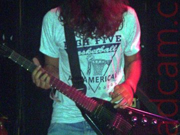 A man with long, shaggy, dark hair, wearing a white t-shirt with a black text logo and a faded blue pair of jeans, and playing a black "flying V" electric guitar, left-handed. His face is hidden in his mop of hair as he looks down at his guitar. Stage lights are giving his t-shirt a blue cast, his jeans a green cast, and his left arm is being high-lighted by a red light. Black walls and ceiling fill the background.