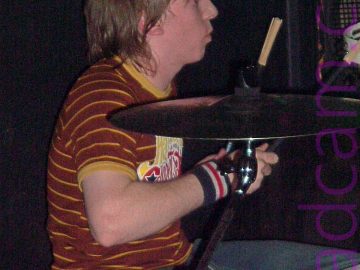 A man with shaggy chin-length brown hair, sitting behind a drum kit facing to the right, with a drum brush raised in his left hand. He is wearing a brown t-shirt with thin yellow bands around the body, and a yellow, white, and red logo on the front. A pair of large cymbals are off to his right, between him and the camera. A large black amplifier is in the background, on the right of the frame, with black walls filling the rest of the frame.
