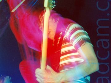A man with shaggy dark hair, wearing a brown and white banded top and a blue pair of jeans, playing a black bass guitar on a stage. A long exposure has combined with the stage lights and his movement to create red and blue light trails tracking where he and his instrument have moved. A woman wearing a sleeveless white top can be seen in the background in the bottom left corner, with black walls and ceiling filling the rest of the frame.