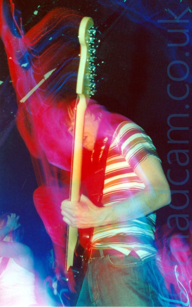 A man with shaggy dark hair, wearing a brown and white banded top and a blue pair of jeans, playing a black bass guitar on a stage. A long exposure has combined with the stage lights and his movement to create red and blue light trails tracking where he and his instrument have moved. A woman wearing a sleeveless white top can be seen in the background in the bottom left corner, with black walls and ceiling filling the rest of the frame.
