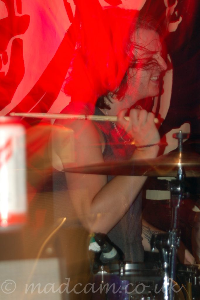 A man with shoulder-length dark hair, wearing a grey vest, sitting behind a sparkly purple drum kit, with sticks held aloft, a grimace on his face, and hair flying everywhere as he rocks out. Red light trails behind him convey a sense of movement.