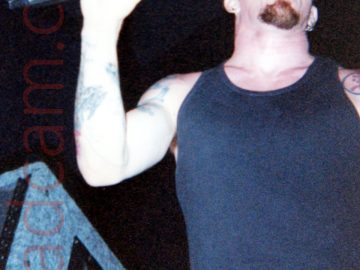 A low shot of a man with a shaved head and a short goatee beard, wearing a black vest top that exposes his heavily tattooed arms, and a blue pair of trousers, performing on stage as part of a rock band. He is holding a microphone in his right hand, the index finger on that hand pointing upwards as he stares into the distance. A large, grey cloth-covered stage speaker is at the side of the stage off to his right, while black walls and ceiling fill the background.