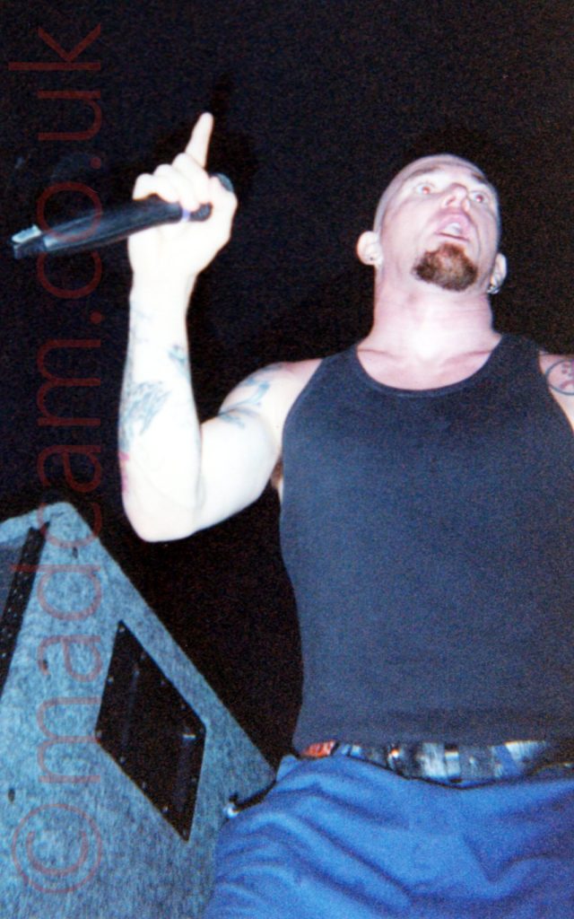 A low shot of a man with a shaved head and a short goatee beard, wearing a black vest top that exposes his heavily tattooed arms, and a blue pair of trousers, performing on stage as part of a rock band. He is holding a microphone in his right hand, the index finger on that hand pointing upwards as he stares into the distance. A large, grey cloth-covered stage speaker is at the side of the stage off to his right, while black walls and ceiling fill the background.