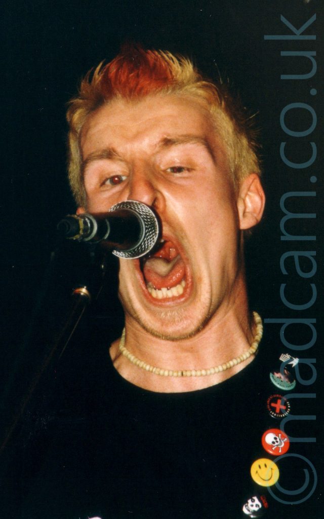 Close-up of a man with short, spikey blond hair, with a red patch right at the front, snarling into a microphone in front of him, his mouth wide open and his eyes partially closed. He has a white-ish beaded necklace round his neck, and is wearing a black t-shirt. A black guitar strap slung over his left shoulder has a row of metal badges - black with a skeletal white face, yellow smiley face, red with a white skull and crossbones, black badge with a red cross and red and white text around the edges, green background with the side of a persons face, black with an anti-littering logo of a person throwing a swastika into a bin. Black walls and ceiling fill the rest of the frame.