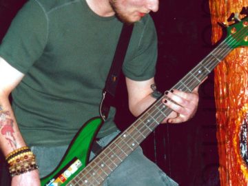 A man with short dark hair and beard, wearing a green t-shirt and a grey pair of cargo pants, playing a bright green 5-stringed bass guitar. His right wrist is covered in an array of beaded bracelet, while his right forearm has a tattoo of a red love heart with a large knife through it. His left forearm has a black tattoo which is partially hidden behind the neck of the bass. He is slightly hunched over, looking down at his left hand. In the background, an orange pole fills the right of the frame, glistening wetly, with a visceral, organic appearance, with black walls and ceiling filling the rest of the frame.