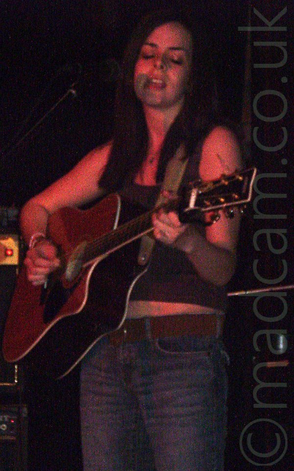 Slightly grainy shot of a woman with long dark hair cascading over her shoulders, wearing a cropped vest-top that exposes her midriff, and a blue pair of jeans with a brown belt, playing a brown acoustic guitar and singing into a microphone on a stand off to the left of the screen. A red light illuminates her, while black walls fill the background.