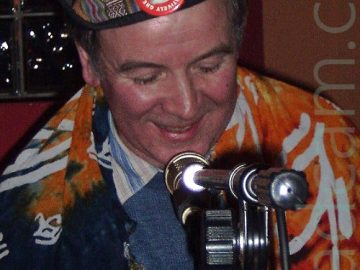 Close-up of the head and shoulders of a man with short, greying hair, wearing a black cap with a multi-coloured, multi-patterned strip of cloth tied around it, surmounted by a red badge with an image of Einstein, and the words "Einstein is relatively great". He is wearing a blue jumper over a blue and white striped shirt, and has a blue and orange bit of cloth draped over his shoulders, with white symbols that could well be an inverted Eye of Horus. A microphone on a stand is in front of him, and the top of a wooden railing is right at the bottom of the frame. The wall behind him is a sort of burnt orange, with red inside around a window made up of fairly small glass bricks.