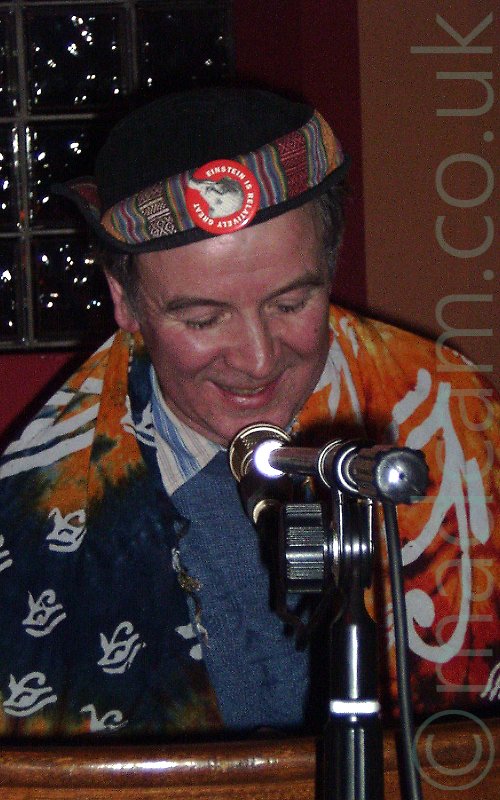 Close-up of the head and shoulders of a man with short, greying hair, wearing a black cap with a multi-coloured, multi-patterned strip of cloth tied around it, surmounted by a red badge with an image of Einstein, and the words "Einstein is relatively great". He is wearing a blue jumper over a blue and white striped shirt, and has a blue and orange bit of cloth draped over his shoulders, with white symbols that could well be an inverted Eye of Horus. A microphone on a stand is in front of him, and the top of a wooden railing is right at the bottom of the frame. The wall behind him is a sort of burnt orange, with red inside around a window made up of fairly small glass bricks.