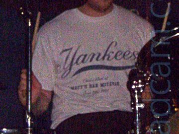 A man with a shaggy little beard, wearing a red woollen hat with wide blue bands, a white t-shirt with a blue "Yankees" logo, and dark trousers, sitting behind a red drum kit, his arms raised slightly into the air, holding his drum sticks. Black walls fill the background.