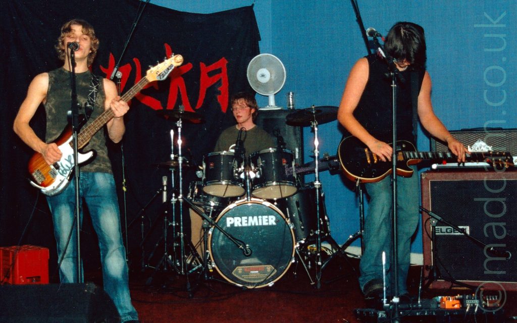 Three young men on a stage, performing as a rock band. On the left is a young man with mid-length wavy blond hair, wearing a green and grey tanktop and a blue pair of jeans, playing a brown and white bass guitar, and singing into a microphone on a stand in front of his face. In the middle is the band's drummer, sitting behind a black drum kit. He has lanky brown hair, and is wearing glasses and a green jumper. A white table-top fan is sitting on top of an amplifier behind him. On the right is a man with mid-length dark hair, wearing a black tanktop and blue jeans, staring down at a black electric guitar he is holding in his hands, almost as if he is wondering how to play it. A black bit of cloth with the word "Tzuka" in red fills the background on the left, with a black and brown amplifier by a blue wall on the right of the frame.