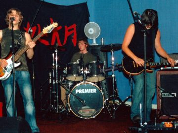Three young men on a stage, performing as a rock band. On the left is a young man with mid-length wavy blond hair, wearing a green and grey tanktop and a blue pair of jeans, playing a brown and white bass guitar, and singing into a microphone on a stand in front of his face. In the middle is the band's drummer, sitting behind a black drum kit. He has lanky brown hair, and is wearing glasses and a green jumper. A white table-top fan is sitting on top of an amplifier behind him. On the right is a man with mid-length dark hair, wearing a black tanktop and blue jeans, staring down at a black electric guitar he is holding in his hands, almost as if he is wondering how to play it. A black bit of cloth with the word "Tzuka" in red fills the background on the left, with a black and brown amplifier by a blue wall on the right of the frame.