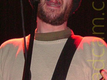 Close-up of a man performing on a stage, singing into a microphone on a stand. He has a short brown beard, wearing a big, black, woolly hat, as well as a beige jumper over a green top. He is playing a black electric guitar, attached to a black strap slung over his shoulder. A red stage light is shining down on him, highlighting his shoulders, head, and left hand. In the background, black walls and white ceiling fill the reast of the frame.