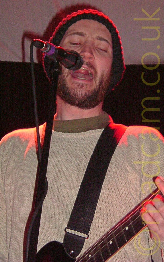 Close-up of a man performing on a stage, singing into a microphone on a stand. He has a short brown beard, wearing a big, black, woolly hat, as well as a beige jumper over a green top. He is playing a black electric guitar, attached to a black strap slung over his shoulder. A red stage light is shining down on him, highlighting his shoulders, head, and left hand. In the background, black walls and white ceiling fill the reast of the frame.