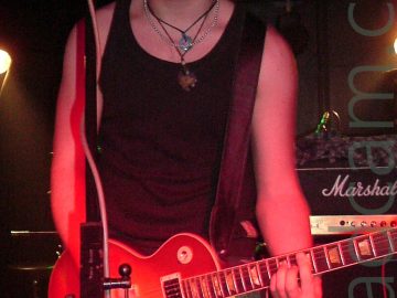 A man with spiky, short, dark hair, and wearing a black vest and dark jeans, playing an orange electric guitar, and looking somewhat tired as he stares at a microphone on a stand in front of him. A black and yellow stripe marks a low ceiling on an alcove behind him, with yellow lights in the alcove illuminating a couple of patches on walls and floor, the rest hidden in darkness.