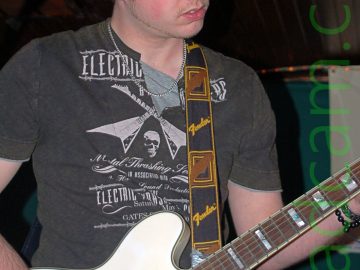 A man wearing a black knitted hat, a black t-shirt with a white print made up of mostly indecipherable text, and a blue pair of jeans. He is playing a white electric guitar, which has a black strap with yellow "Fender" text and a brown representation of a guitar head slung over his left shoulder. A microphone on a stand is in front of his head up in the top left corner. Gren walls and a brown wooden ceiling fill the background.