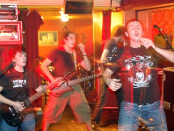 Four of the five members of a band putting on a rather energetic performance on a stage. On the left is a fairly short young man with dark hair, wearing a black t-shirt with a white "The Idiot Rate" logo emblazoned across the chest, and playing a black electric guitar. In the middle is a man with short dark hair, weaing a plain black t-shirt and abeige pair of knee-length shorts, playing a brown electric guitar, with a look of surprise on his face. On the right is the bands singer, with short dark hair, wearing a black t-shirt with a white and red logo featuring a skull, dragons, and a pegasus, on the front. He is holding a microphone in his left hand, and is conspicuously not singing into it right now, but that is all about to change. Behind him is a tall man with shaggy brown hair and a brown beard, wearing a grey t-shirt, and playing a bass guitar. Orange and brown walls almost look golden in the yellow room lights, with red curtains hangin loose on some of the walls. Stacks of amps and monitors line some of the walls behind the band. A long exposure combines with the bands movement to create streaks of red light around them, along with secondary images through which the background can be seen.