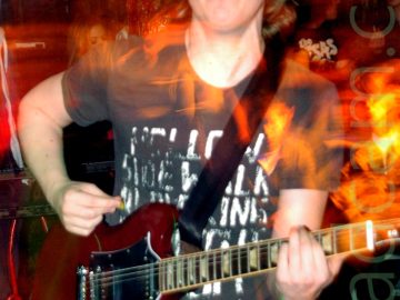 A man with floppy, shoulder-length blond hair, wearing a black t-shirt with faded white text filling the front, and a blue pair of jeans, playing a red electric guitar. An orange pole stretches from floor to ceiling behind him, painted to look sticky and organic and visceral, with some people beyond that, with dark coloured walls behind them. A long exposure and his movement have created red and orange streaks of light and secondary images.