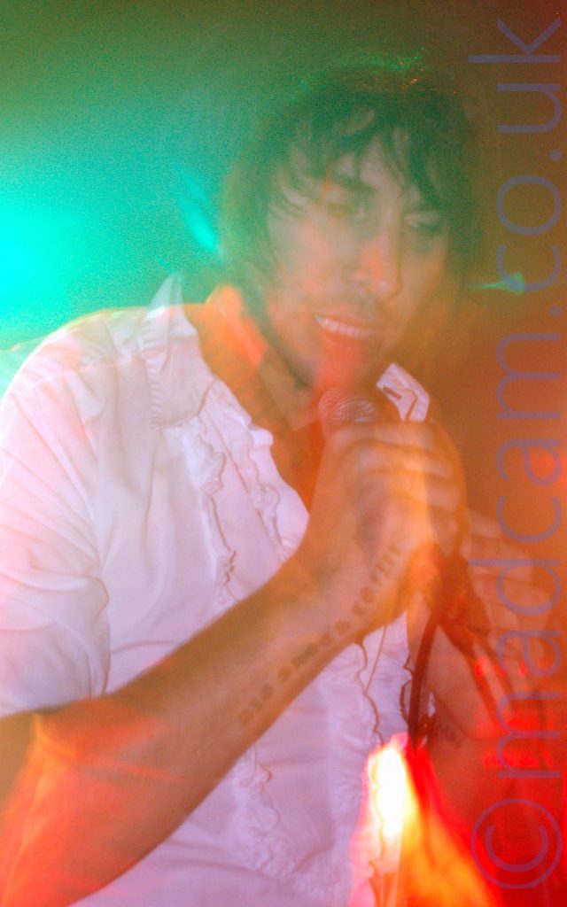 Close-up of a man with shaggy dark hair, wearing a frilly white shirt, singing into a microphone held in his right hand. The words "Big shoes to fill" have been tattooed on the inside edge of his right forearm. A long exposure has made him slightly indistinct, his movement and blooms of red and green lights creating artifacts and secondary images all over the frame.
