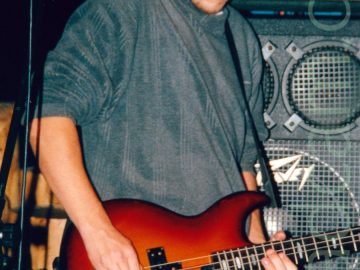 A man with short dark hair and long sideburns, wearing a grey jumper and beige trousers, and playing a red electric bass guitar. A microphone on a stand is in front of his right arm. A wall of grey and black amplifiers is stacked behind him.