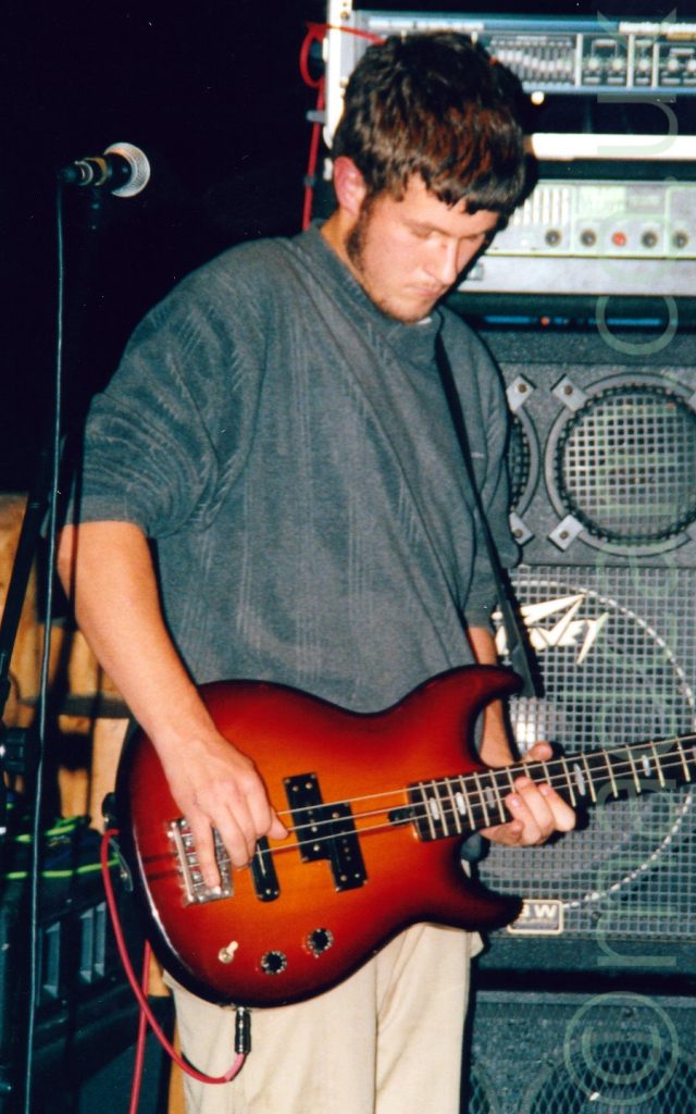A man with short dark hair and long sideburns, wearing a grey jumper and beige trousers, and playing a red electric bass guitar. A microphone on a stand is in front of his right arm. A wall of grey and black amplifiers is stacked behind him.